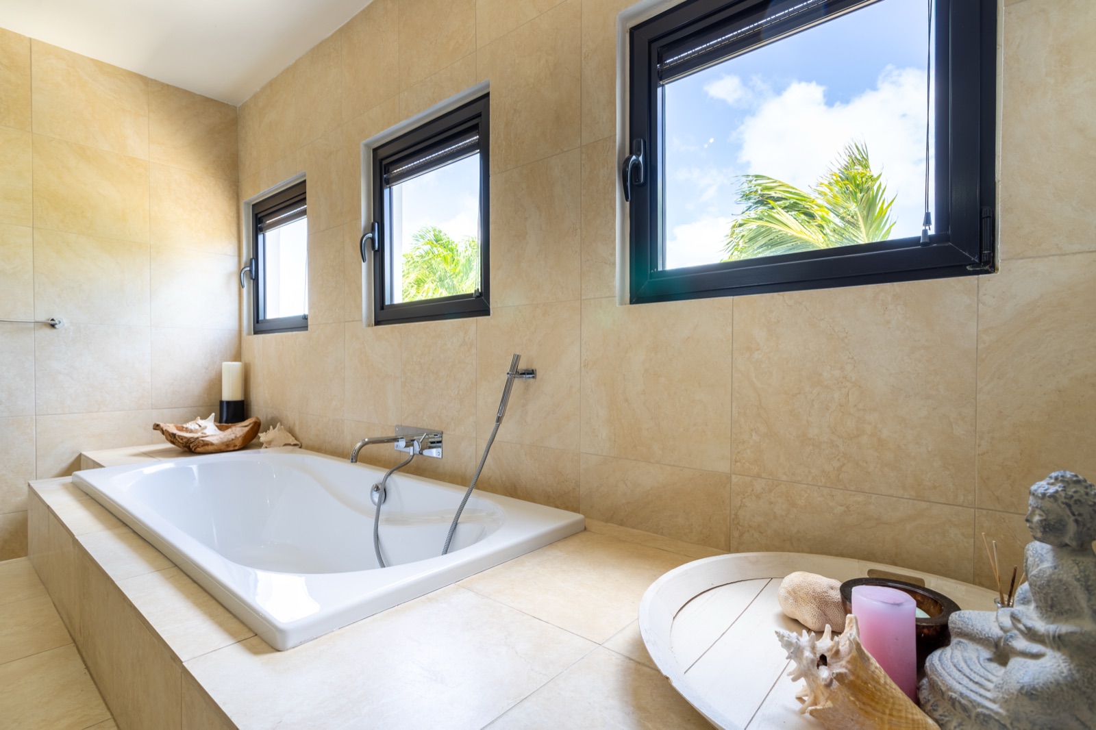 Luxury Bonaire villa spa bathroom with a deep soaking tub, natural stone tile, and windows showing a view of blue sky and palm trees.